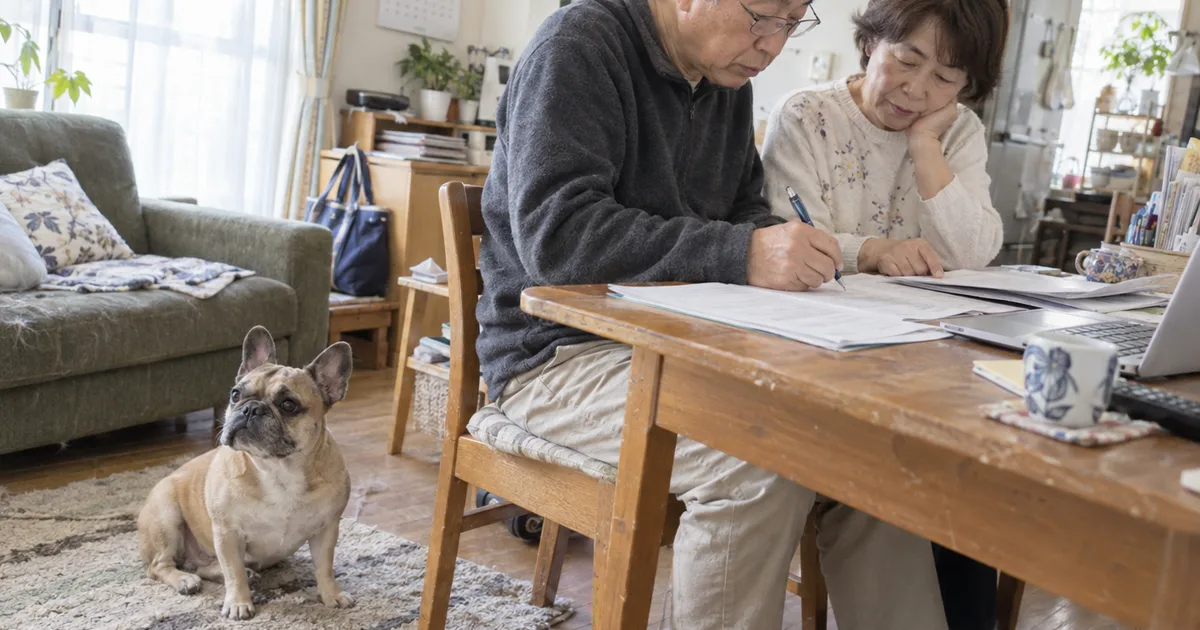 自宅の机で書類に記入する60代の夫婦と、足元に座るフレンチブルドッグ