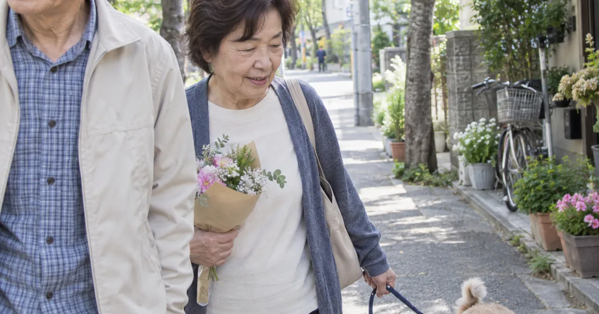 花束を持ち、リードをつけた小型犬と並木道を歩く60代の夫婦